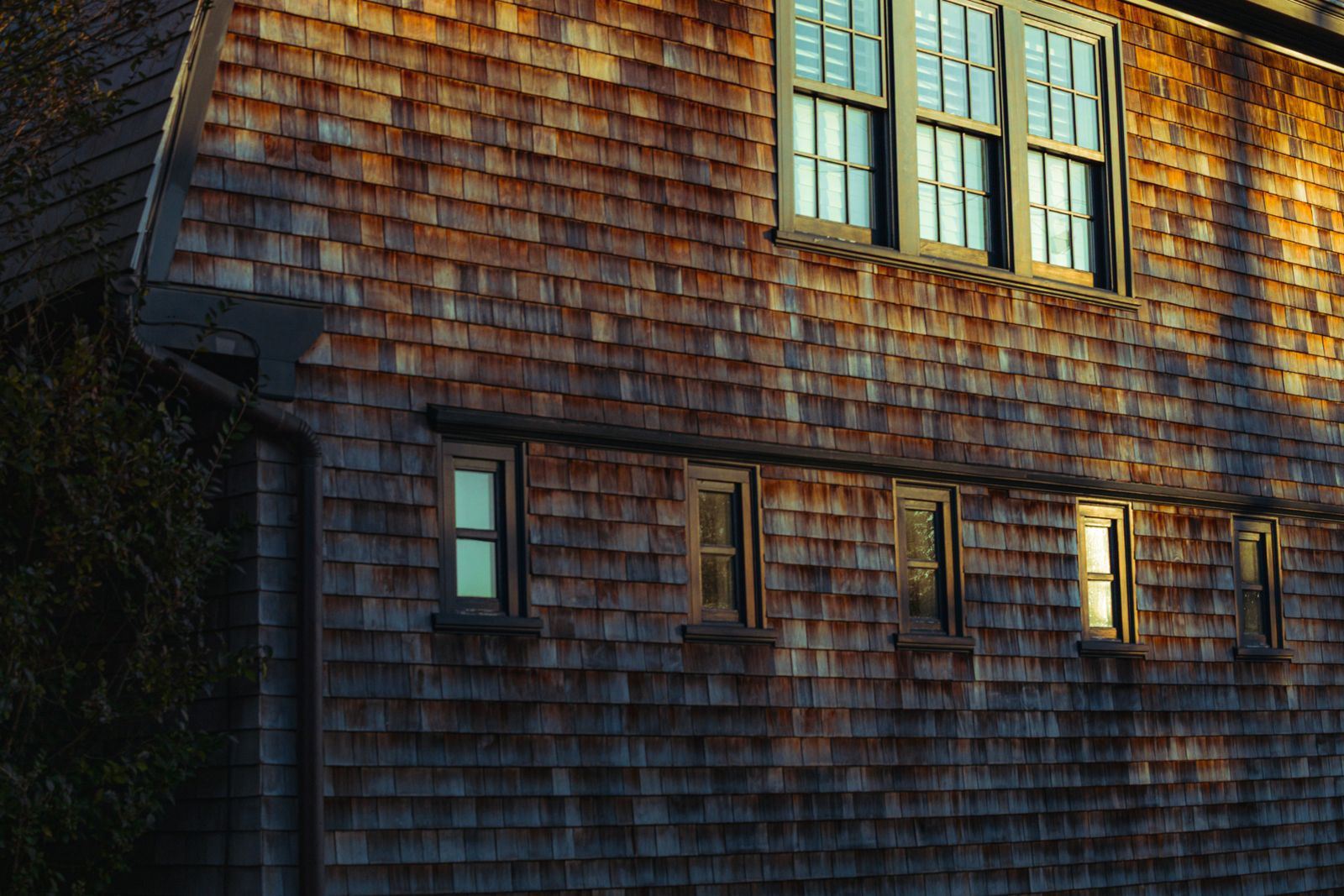 A two-storey house clad in weathered cedar shingles with white-trimmed windows, in warm evening light